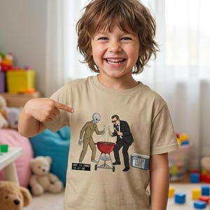 Puede incluir: Una camiseta color beige con una ilustración de dibujos animados de un extraterrestre y un hombre con traje haciendo una barbacoa. El diseño incluye una barbacoa roja, un código de barras y texto. El niño sonríe y señala la camiseta.