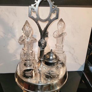 May include: A vintage silver-plated condiment set with four clear glass bottles and jars. The set includes a central handle with an ornate design and a round base. The glass containers have decorative stoppers and lids.