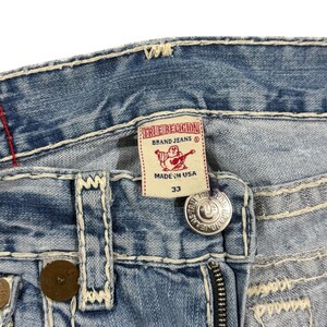 May include: Light blue denim jeans featuring thick white contrast stitching, a metal button closure, and a white interior label with red text reading True Religion Brand Jeans, a red Buddha logo, and the size 33, all made in the USA.