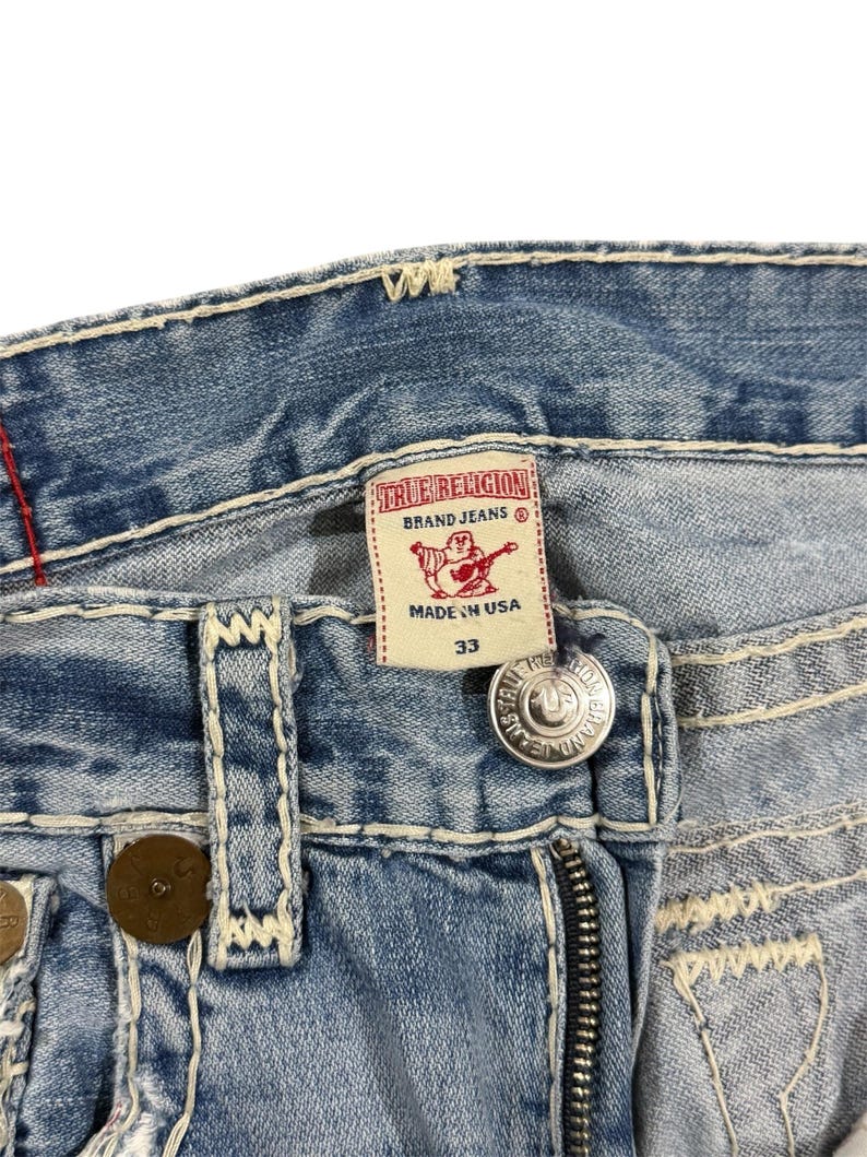 May include: Light blue denim jeans featuring thick white contrast stitching, a metal button closure, and a white interior label with red text reading True Religion Brand Jeans, a red Buddha logo, and the size 33, all made in the USA.