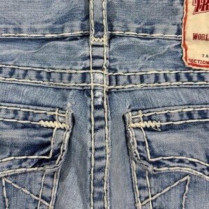 May include: Light blue denim jeans featuring prominent thick white contrast stitching, two back pockets with flap closures, and a rectangular brand label in the upper right corner with the visible text TR, WORLD, and SECTION.