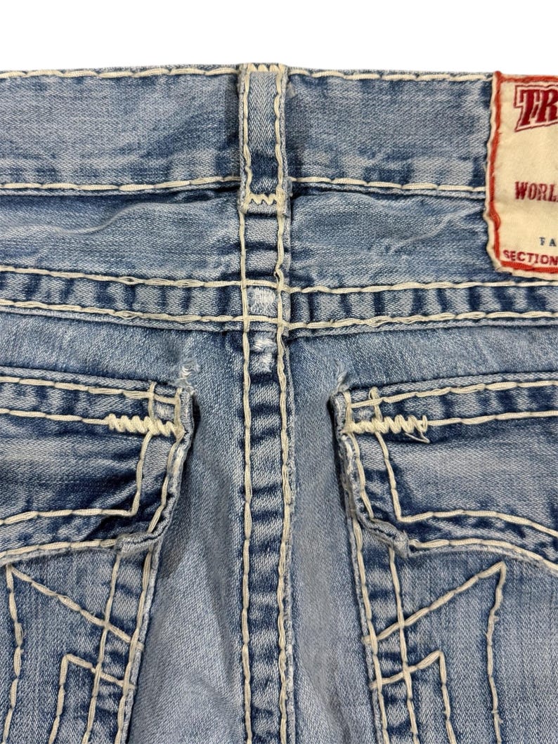 May include: Light blue denim jeans featuring prominent thick white contrast stitching, two back pockets with flap closures, and a rectangular brand label in the upper right corner with the visible text TR, WORLD, and SECTION.