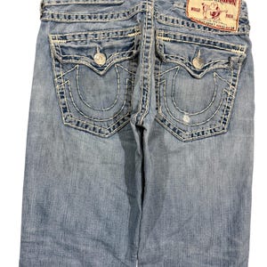 May include: Light blue denim jeans featuring thick white contrast stitching, two back flap pockets with silver-tone button closures, and a red True Religion brand patch on the waistband. The jeans display a faded wash and a small distressed area.