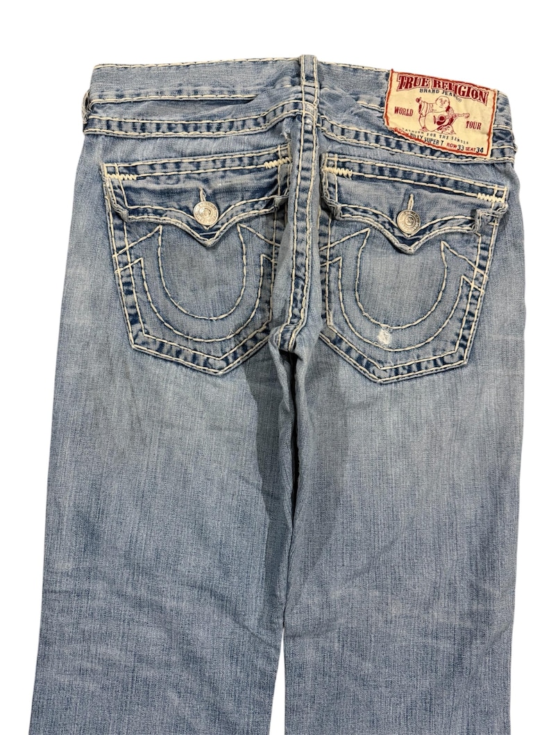 May include: Light blue denim jeans featuring thick white contrast stitching, two back flap pockets with silver-tone button closures, and a red True Religion brand patch on the waistband. The jeans display a faded wash and a small distressed area.