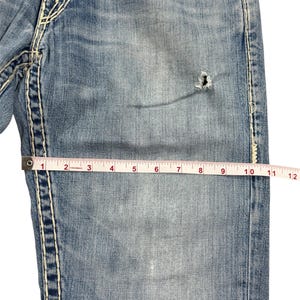 May include: Light blue denim jeans featuring thick white contrast stitching, a small distressed hole on the thigh, and a metal button. A measuring tape placed across the fabric shows a width of approximately 12 inches at this specific point.