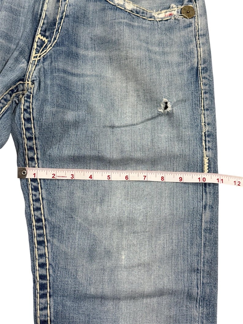 May include: Light blue denim jeans featuring thick white contrast stitching, a small distressed hole on the thigh, and a metal button. A measuring tape placed across the fabric shows a width of approximately 12 inches at this specific point.