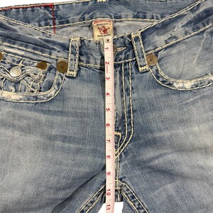 May include: Light blue denim jeans with heavy white contrast stitching, distressed detailing, and metallic rivets. A white measuring tape is placed vertically over the center, showing a rise measurement of approximately 13 inches from the crotch seam.