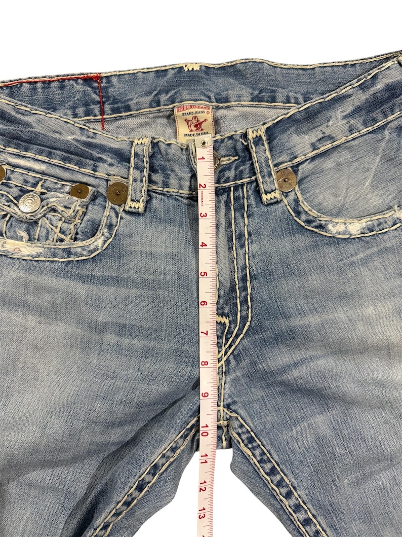 May include: Light blue denim jeans with heavy white contrast stitching, distressed detailing, and metallic rivets. A white measuring tape is placed vertically over the center, showing a rise measurement of approximately 13 inches from the crotch seam.