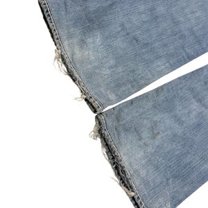 May include: A pair of light blue denim jeans featuring a faded wash and distressed, frayed hems at the bottom of both pant legs. The fabric shows a classic twill texture with subtle whiskering and wear patterns consistent with well-worn denim.