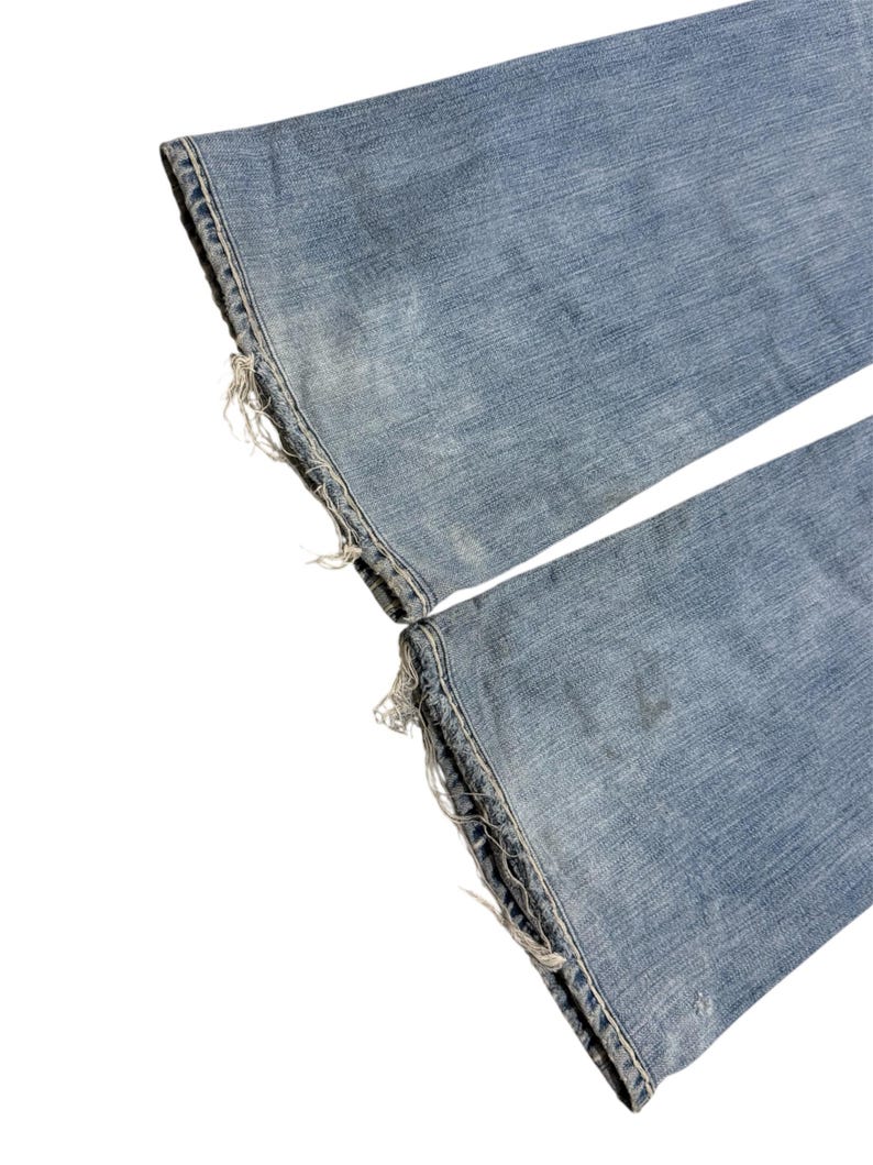 May include: A pair of light blue denim jeans featuring a faded wash and distressed, frayed hems at the bottom of both pant legs. The fabric shows a classic twill texture with subtle whiskering and wear patterns consistent with well-worn denim.