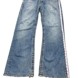 May include: Light blue denim jeans with heavy contrast stitching, featuring a distressed finish, button fly, and front pockets. A white measuring tape is placed along the side, showing a total length of approximately 42 inches from waist to hem.