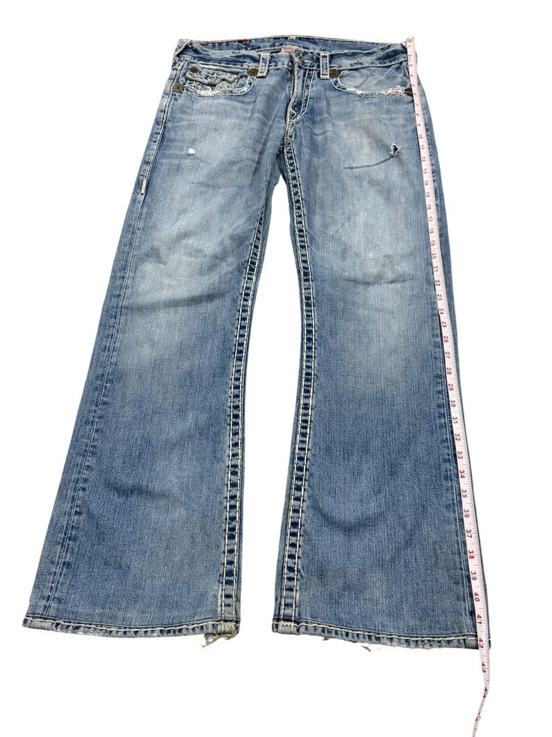 May include: Light blue denim jeans with heavy contrast stitching, featuring a distressed finish, button fly, and front pockets. A white measuring tape is placed along the side, showing a total length of approximately 42 inches from waist to hem.