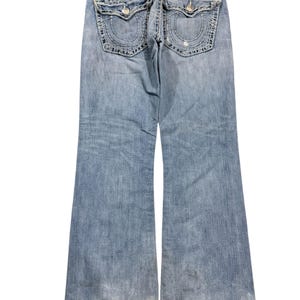 May include: Light blue denim jeans featuring a back view with two flap pockets, thick white contrast stitching, and a True Religion brand patch on the waistband. The jeans have a bootcut leg silhouette with frayed hems and a faded wash finish.