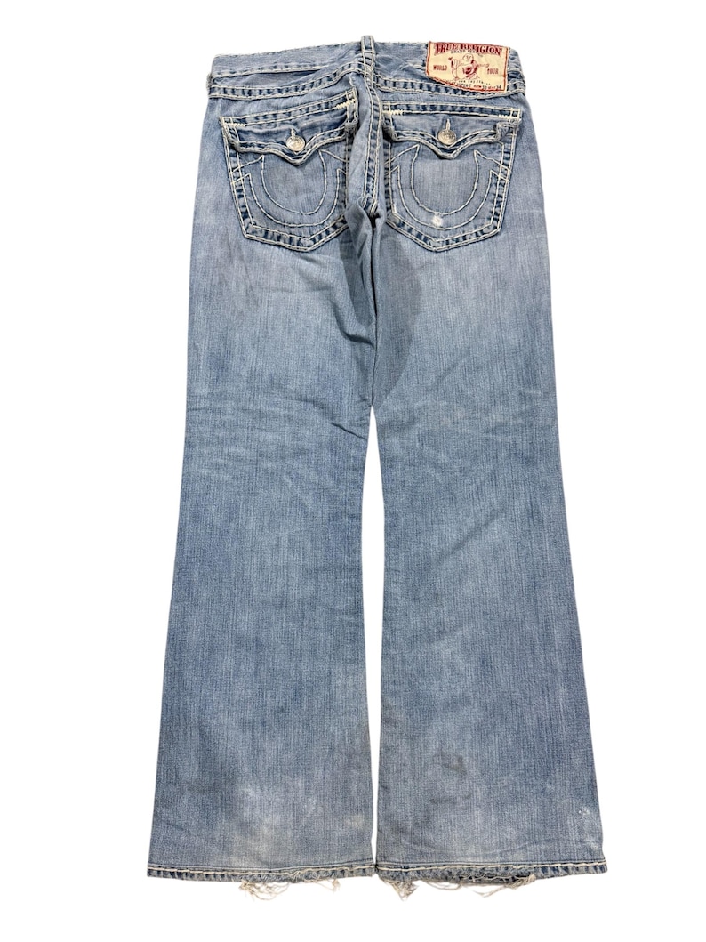 May include: Light blue denim jeans featuring a back view with two flap pockets, thick white contrast stitching, and a True Religion brand patch on the waistband. The jeans have a bootcut leg silhouette with frayed hems and a faded wash finish.