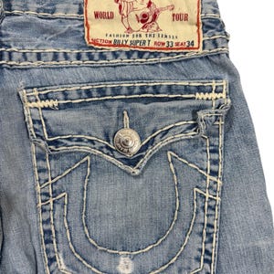 May include: Light blue denim jeans featuring thick white contrast stitching, a back pocket with a button flap, and a prominent horseshoe-shaped stitch design. A rectangular patch at the waist displays the text True Religion Brand Jeans.