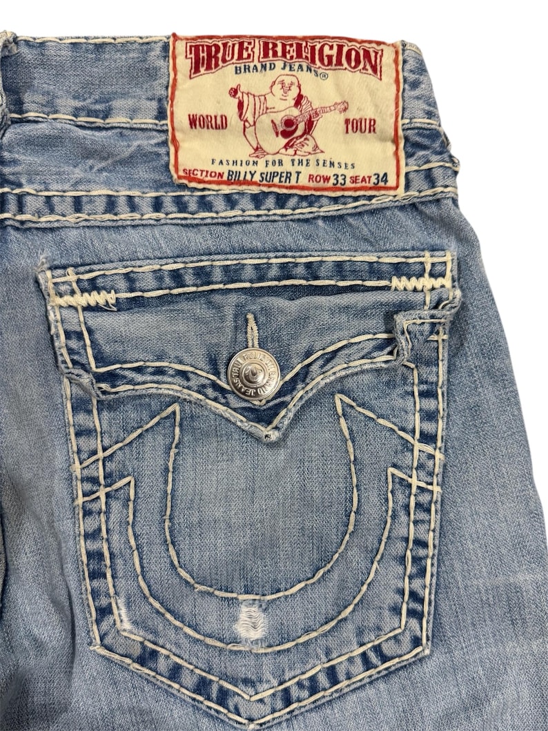 May include: Light blue denim jeans featuring thick white contrast stitching, a back pocket with a button flap, and a prominent horseshoe-shaped stitch design. A rectangular patch at the waist displays the text True Religion Brand Jeans.
