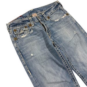 May include: Light blue denim jeans featuring a faded wash, distressed details, and prominent thick white contrast stitching. The jeans include a coin pocket, metal rivets, a button fly closure, and a branded label on the interior waistband.