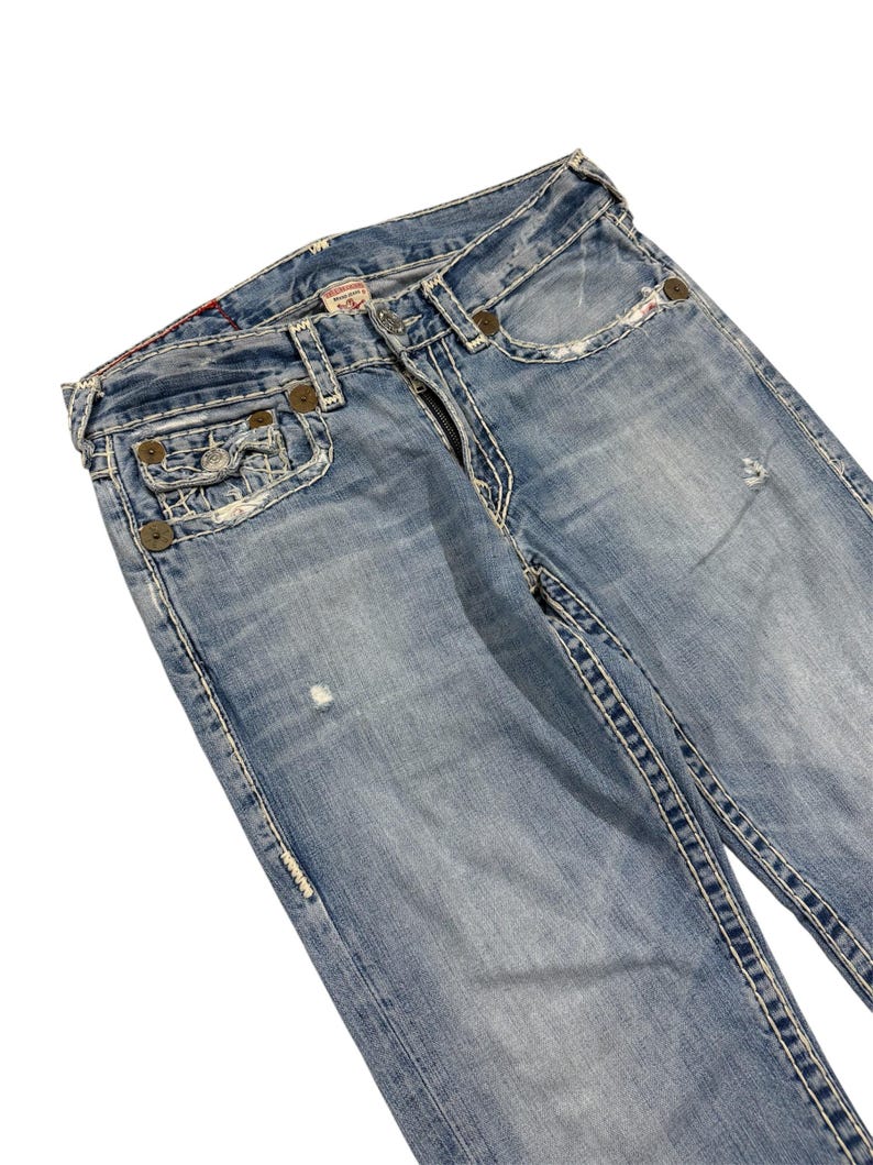 May include: Light blue denim jeans featuring a faded wash, distressed details, and prominent thick white contrast stitching. The jeans include a coin pocket, metal rivets, a button fly closure, and a branded label on the interior waistband.