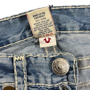 May include: Light blue denim jeans featuring thick, cream-colored contrast stitching, a silver-toned metal button embossed with True Religion branding, and a white interior care label with black text detailing 100% cotton composition.