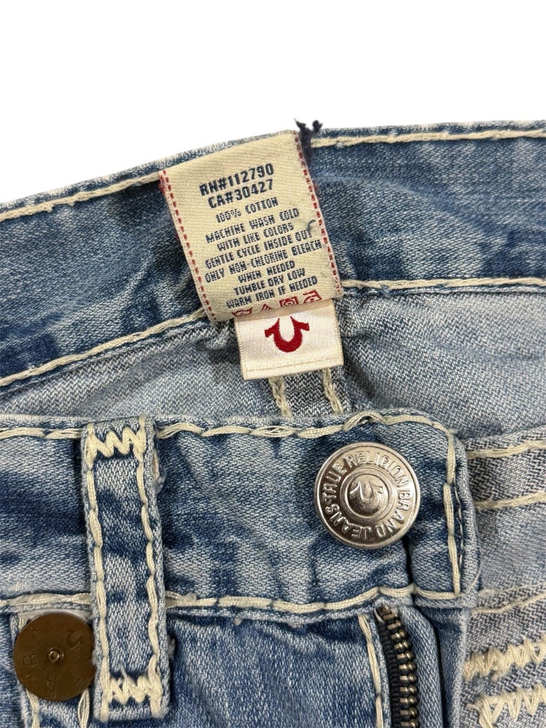 May include: Light blue denim jeans featuring thick, cream-colored contrast stitching, a silver-toned metal button embossed with True Religion branding, and a white interior care label with black text detailing 100% cotton composition.