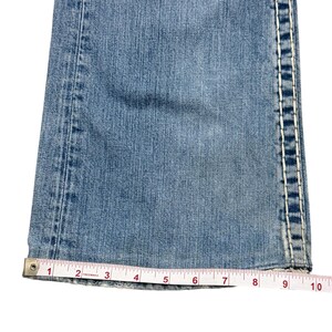 May include: Light blue denim jeans leg featuring thick white contrast stitching along the side seams. A white measuring tape is positioned across the hem, showing a width measurement of approximately 10 inches at the bottom of the pant leg.