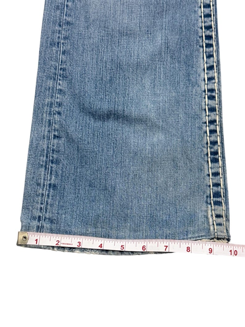May include: Light blue denim jeans leg featuring thick white contrast stitching along the side seams. A white measuring tape is positioned across the hem, showing a width measurement of approximately 10 inches at the bottom of the pant leg.