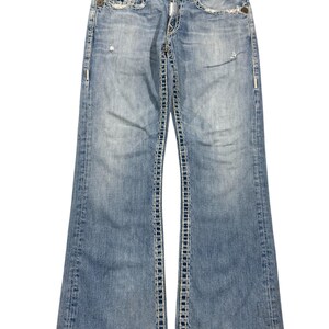 May include: Light blue denim jeans featuring a distressed finish, a button fly, and prominent thick white contrast stitching along the seams and pockets. The jeans have a bootcut leg silhouette with fading on the thighs and knees.