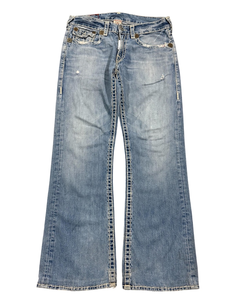 May include: Light blue denim jeans featuring a distressed finish, a button fly, and prominent thick white contrast stitching along the seams and pockets. The jeans have a bootcut leg silhouette with fading on the thighs and knees.