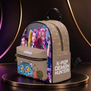 May include: A tan backpack featuring a graphic print of characters from K-Pop Demon Hunters, a blue cat illustration, and the text HUNTRIX and K-POP DEMON HUNTERS. The bag has a top handle, black zippers, and a repeating monogram pattern on the side.