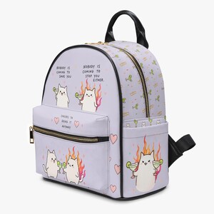 Könnte beinhalten: Ein hellvioletter Rucksack mit Cartoon-Katzen, Margaritas und Flammen. Der Text lautet: "Nobody is coming to save you. Nobody is coming to stop you either." und "Cheers to doing it anyway." Der Rucksack hat schwarze Ränder und goldene Reißverschlüsse.