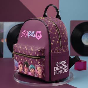 May include: A purple backpack featuring a repeating gold pattern of stars and diamonds. The front pocket displays four illustrated characters, while the top section reads "Saja Boys" and the side panel reads "K-Pop Demon Hunters" in white text.