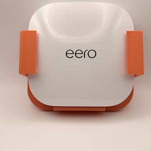May include: A white and orange eero Wi-Fi router. The router has a rounded rectangular shape with the brand name "eero" printed in black. It features two orange brackets on the sides.