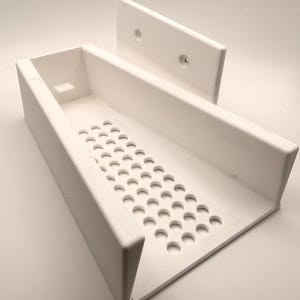May include: A white, rectangular, 3D-printed tray with a perforated base and a separate mounting piece. The tray has a raised edge and a small rectangular cutout. The mounting piece has two holes for attachment.