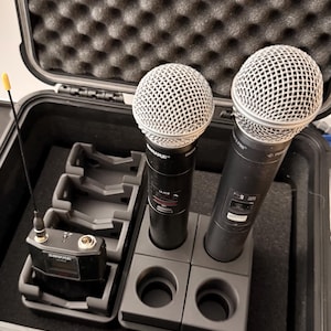 Può includere: Una valigetta rigida nera con imbottitura in schiuma contiene due microfoni wireless e un ricevitore. I microfoni hanno testine a rete argentate e corpi neri. La valigetta contiene anche basi di ricarica.