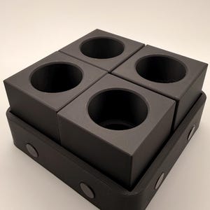 Puede incluir: Un organizador negro de forma cuadrada con cuatro aberturas circulares, colocado sobre una base rectangular. El organizador est&aacute; hecho de un material mate y tiene un dise&ntilde;o minimalista. La base tiene esquinas redondeadas y peque&ntilde;os acentos circulares.