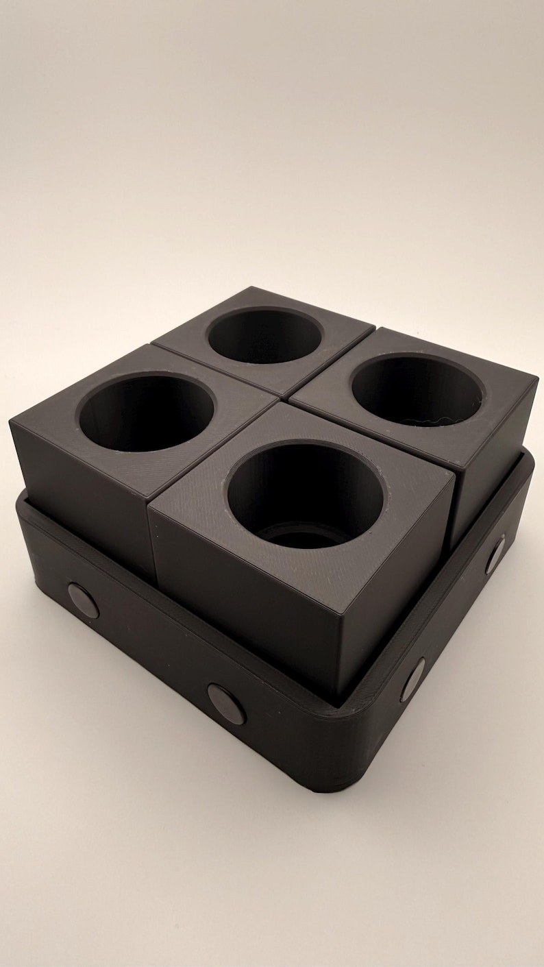 Puede incluir: Un organizador negro de forma cuadrada con cuatro aberturas circulares, colocado sobre una base rectangular. El organizador est&aacute; hecho de un material mate y tiene un dise&ntilde;o minimalista. La base tiene esquinas redondeadas y peque&ntilde;os acentos circulares.