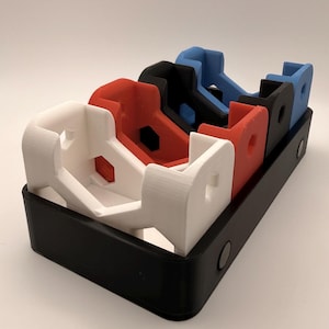 Puede incluir: Una bandeja negra contiene cuatro objetos impresos en 3D en blanco, rojo, negro y azul. Cada objeto tiene una forma distintiva, con una base rectangular y una parte superior curva.