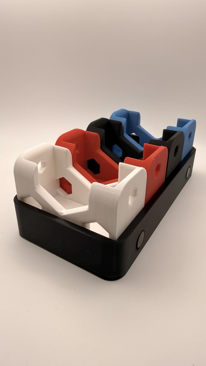 Puede incluir: Una bandeja negra contiene cuatro objetos impresos en 3D en blanco, rojo, negro y azul. Cada objeto tiene una forma distintiva, con una base rectangular y una parte superior curva.