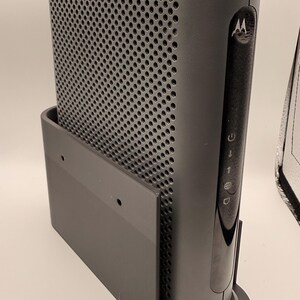 Puede incluir: Un módem-router gris oscuro con un panel frontal perforado y un panel lateral negro brillante curvado. El dispositivo está montado en un soporte gris a juego. El logotipo de Motorola es visible en el lateral.