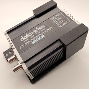 May include: A black Datavideo UP/DOWN CROSS CONVERTER DAC-70. The device has silver-colored connectors and the text "datavideo" and "www.datavideo.com" printed on the top. It also has a mode select panel with various options.