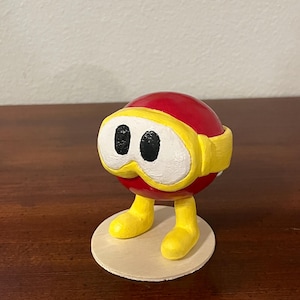 Puede incluir: Una figura pintada a mano de una esfera roja con gafas y piernas amarillas, de pie sobre una base de madera redonda. La figura tiene ojos negros y está pintada con colores primarios brillantes. El diseño recuerda a un personaje de dibujos animados.