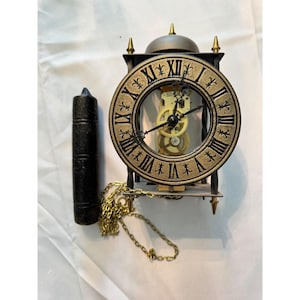 May include: An antique clock with a gold-colored face and black Roman numerals. The clock has a visible mechanical movement and is encased in a black frame. A black handle and gold chain are also present.