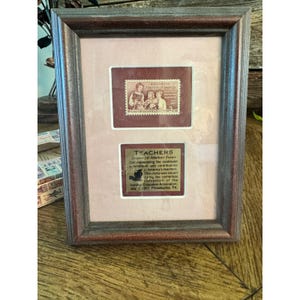 May include: Framed vintage stamp art honoring American teachers. The artwork features a maroon-colored stamp with the text "Honoring the Teachers of America" and a commemorative plaque below. The frame is a dark brown color.