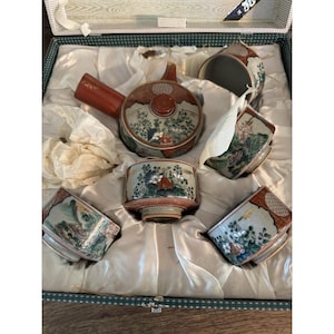 Op de afbeelding: Een vintage Japanse theeset in een doos. De set bevat een theepot met een rode handgreep en deksel, en vier bijpassende kopjes. De porseleinen stukken zijn versierd met een landschapsscène in tinten groen, rood en blauw.