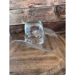 May include: A clear glass sculpture shaped like a cowboy hat. The object features a rounded crown and a wide, curved brim, all crafted from transparent, smooth glass. The design mimics the classic silhouette of a western-style hat in glass.