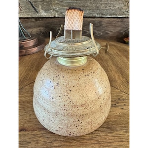 May include: A vintage oil lamp with a speckled, beige ceramic base and a brass-colored metal top. The top features the word "EAGLE" and a wick. The lamp is designed for use with oil and has a classic, rustic aesthetic.