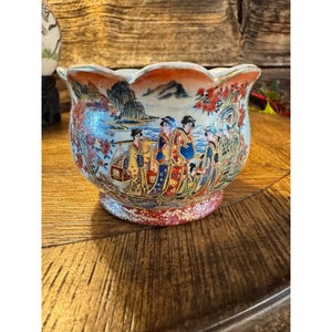 May include: A decorative ceramic bowl with a scalloped rim. The bowl features a detailed, colorful illustration of figures in traditional attire, set against a landscape with mountains and water. The base has a red floral pattern.