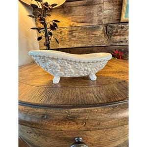 May include: A small, white ceramic bathtub with floral detailing. The decorative tub has three small feet and a scalloped edge. The tub is displayed on a wooden surface.