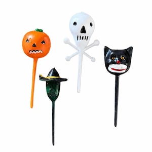 May include: A set of four Halloween-themed decorative picks featuring an orange pumpkin, a white skull with crossbones, a black cat face, and a green figure wearing a black witch hat. Each figure is attached to a thin, straight stem for insertion.