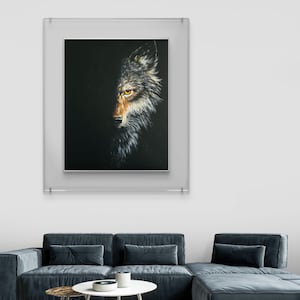 Puede incluir: Obra de arte enmarcada que presenta una pintura detallada de la cara de un lobo emergiendo de la oscuridad. El ojo del lobo es de un amarillo llamativo, y su pelaje está representado en tonos grises, marrones y blancos. La obra se exhibe en un salón moderno.